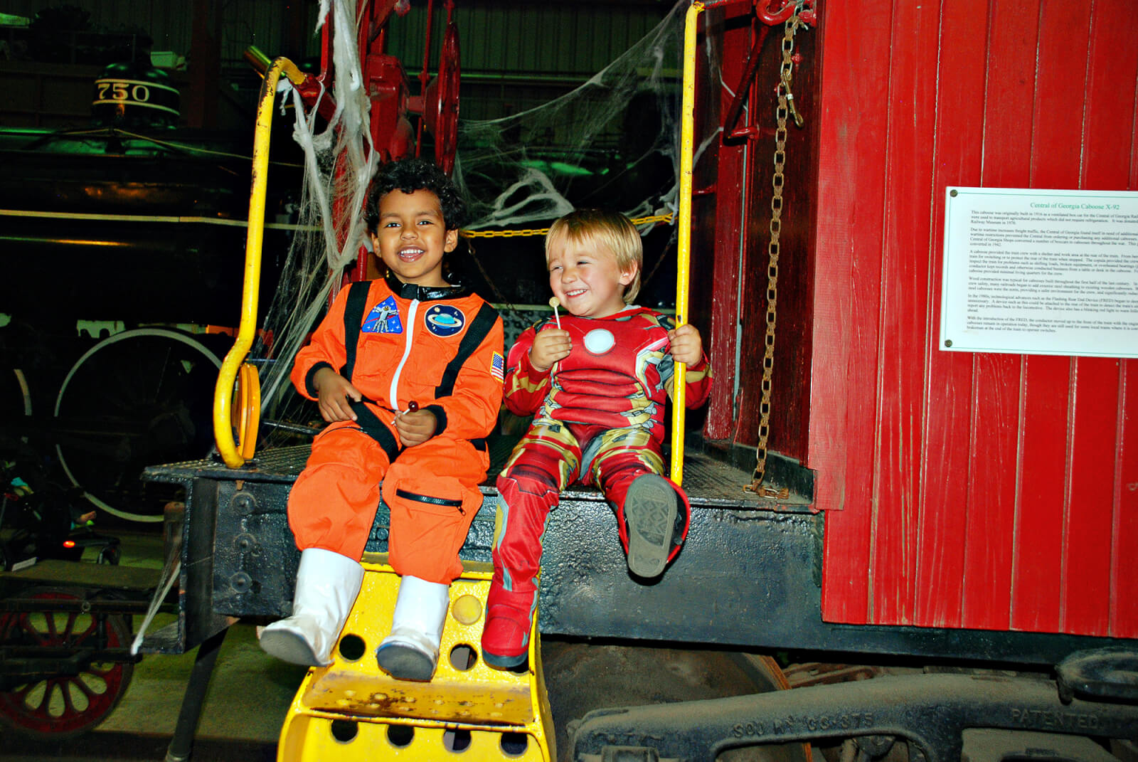 Two boys sit and laugh on the end of a train caboose, while dressed in costumes and eating candy, at Train or Treat - an annual Halloween event at Southeastern Railway Museum. 